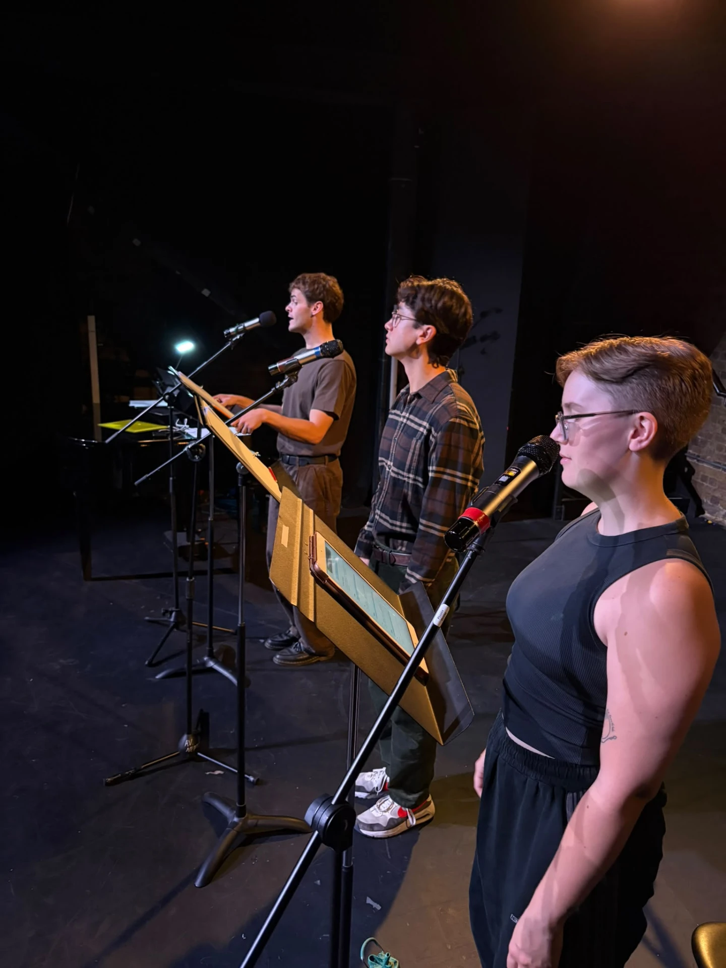 Three performers in rehearsal, standing at microphones and reading from scripts on music stands.
