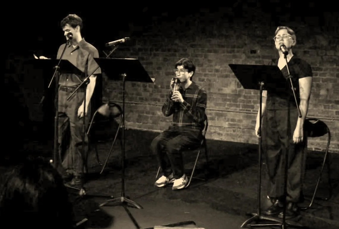 Josh Liew as Wilfred Owen seated and playing a recorder, with Charlie Renwick as Siegfried Sassoon on the left and Maddie Gray as the Witness singing on the right.