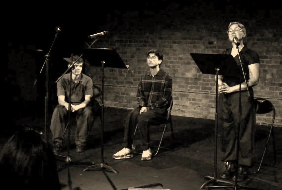 Maddie Gray as the Witness singing at a microphone, with Charlie Renwick as Siegfried Sassoon and Josh Liew as Wilfred Owen seated beside her.