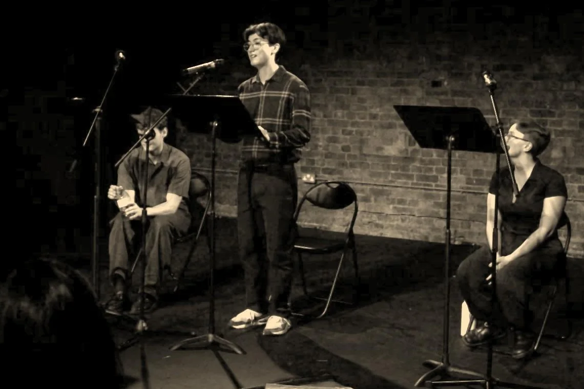 Josh Liew as Wilfred Owen standing at a microphone, with Charlie Renwick as Siegfried Sassoon seated on the left and Maddie Gray as the Witness seated on the right.